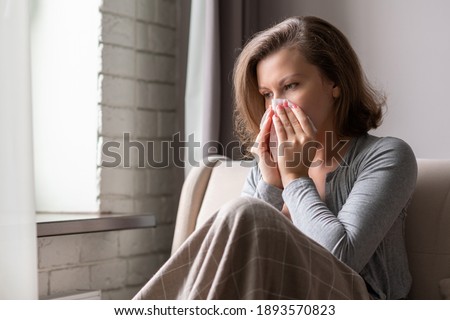 Sick brunette woman sneezing in a tissue sitting on the couch in the living room near window