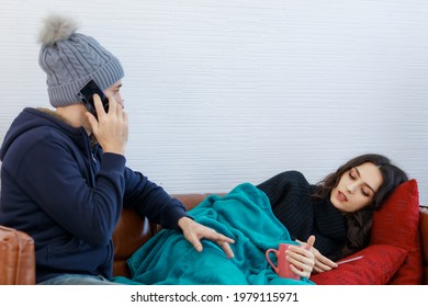Sick Caucasian Female In Wool Sweater Lay Down On Pillow And Leather Sofa Cover By Blanket Having Flu Symptoms In Winter While Husband In Jacket And Knitted Hat Call Doctor At Hospital For Advice.