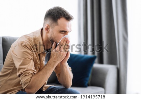 Sick caucasian guy sits on couch feel unhealthy, he suffering from rhinitis snuffles, having respiratory infection, get flue, sitting at home at quarantine