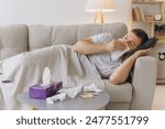 Sick caucasian man with a runny nose lying under a warm blanket on the sofa at home and sneezing in paper tissue. Cold, flu, seasonal winter infections concept