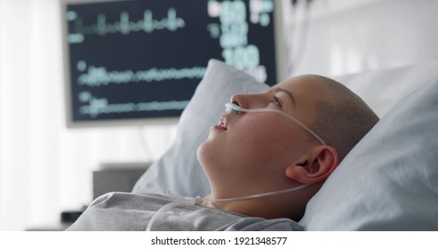 Sick Child With Cancer Lying In Hospital Bed With Nasal Cannula. Close Up Portrait Of Bald Teen Boy Patient Resting In Bed After Chemotherapy Treatment Or Surgery