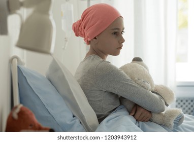 Sick Child With Cancer Sitting In Hospital Bed Holding Teddy Bear