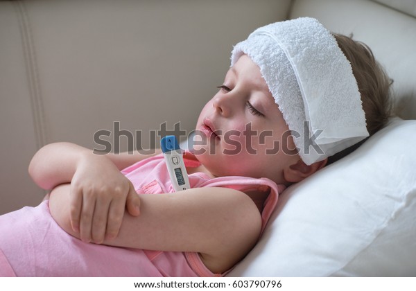  Sick child with high fever laying\
in bed and  holding thermometer.  Compress on\
forehead.