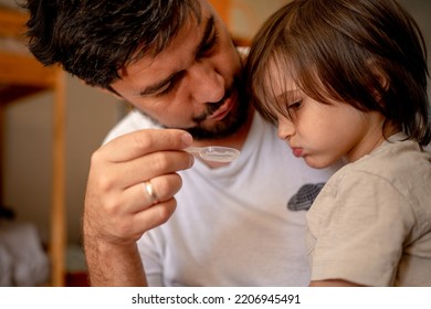 A Sick Child Is Sitting On His Father's Lap. Dad Tries To Get His Son Drunk With Medicine