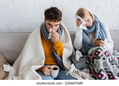 Sick Couple Covered In Blankets Holding Cups Of Tea And Tissues While Sitting On Sofa