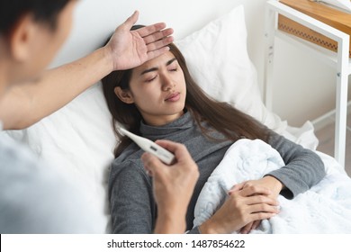 Sick Day At Home In Winter Season. Young Asian Woman Lying On The Bed Have A Fever And Her Husband Checking Thermometer And Measure Body Temperature.