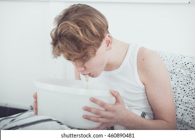 Sick or drunk young boy vomiting into a white bowl as he sits on his bed at home, close up realistic view