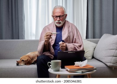 Sick elderly man checking his temperature suffering from seasonal flu or cold and using digital tablet for entertainment or online doctor consultation. Ill senior feel unhealthy with influenza at home - Powered by Shutterstock