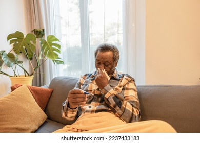 Sick elderly man checking his temperature because of seasonal flu or cold looks at his temperature with thermometer. Sick African American elderly man with flu symptoms at home. - Powered by Shutterstock