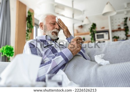 Sick elderly man checking his temperature suffering from seasonal flu or cold lying on sofa caughing suffering from seasonal flu or cold. . Ill senior feel unhealthy with influenza at home