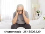 Sick elderly man is suffering from a headache or migraine while sitting on bed at home. Health problems, with the man feeling stressed, sad, and in pain, indicating his discomfort.