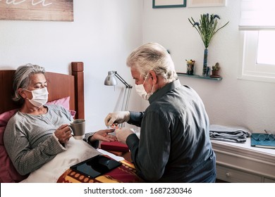 Sick elderly woman of COVID-19 lies in bed at home wearing medical mask. Her husband takes care of her. Two senior people at home. Quarantine self-isolation concept - Shutterstock ID 1687230346