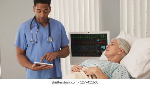 Sick Elderly Woman In Hospital Bed Connected To ECG Monitor Talking To Nurse