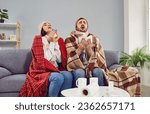 Sick family couple wrapped in blankets sneezing in paper tissues at home. Freezing man and woman in hats wrapped plaids suffering from common cold, flu sitting on couch. Seasonal respiratory diseases