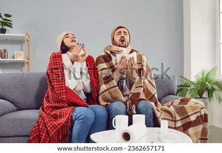 Sick family couple wrapped in blankets sneezing in paper tissues at home. Freezing man and woman in hats wrapped plaids suffering from common cold, flu sitting on couch. Seasonal respiratory diseases