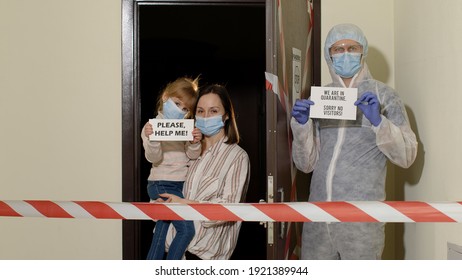 Sick Family Of Mother And Daughter Opening Door With Warning Tapes In Corridor Entrance For Doctor In PPE Biohazard Suit With Slogan In Hands. Visiting Patient At Home. Coronavirus Quarantine Lockdown