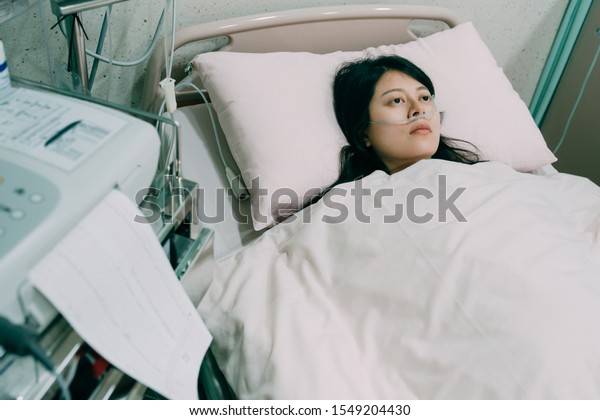 Sick female patient lying on bed wear nasal\
cannula. illness asian chinese woman cover body with white sheet in\
hospital room. upset lady with cancer depressed looking at ceiling\
with machine beside