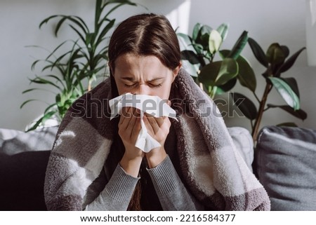 Sick frozen young female seated on sofa in living room covered with warm cozy plaid sneezing holding paper napkin blow out runny nose feels unhealthy, seasonal cold. Weakened immune system concept.
