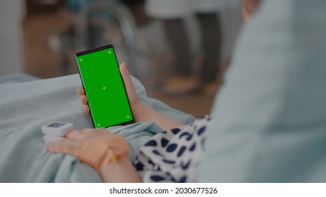 Sick Girl Holding Mock Up Green Screen Chroma Key Phone With Isolated Display During Recovery Consultation In Hospital Ward. Child With Oximeter On Finger Resting In Bed After Suffering Medical