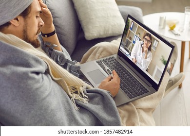Sick Guy At An Online Consultation. Man Looks At A Thermometer Talking To His Doctor On A Video Call While Staying At Home. Man Wrapped In A Plaid Consults During An Online Visit To The Doctor.