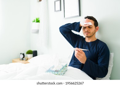 Sick Hispanic Man With Fever And The Flu Checking His Temperature While Resting In Bed During The Morning 