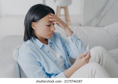 Sick Hispanic Young Woman Is Measuring Temperature At Home. Girl Is Sitting On Sofa And Looking At The Display Of Electronic Thermometer. Headache From Coronavirus Or Influenza. Sick Leave Concept.