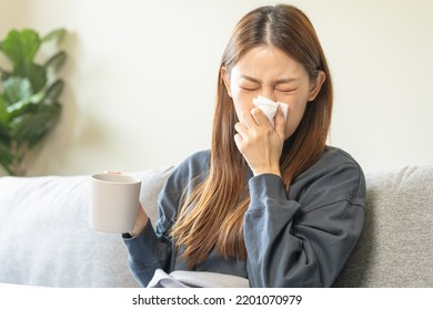 Sick, Hurt Or Pain Asian Young Woman Have A Fever, Flu Sore Throat, Drinking A Mug Of Warm Water, Use Tissues Paper Sneezing Nose, Runny While Sitting Rest On Sofa At Home. Health Care On Virus Person