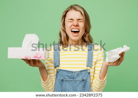 Sick ill allergic woman has red watery eyes runny stuffy sore nose suffering from allergy trigger symptoms hay fever holding paper napkin handkerchief sneeze isolated on plain green background studio