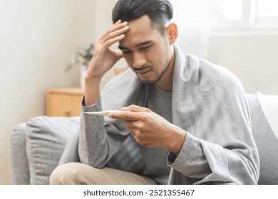 Sick, influenza asian young man, male have fever hand holding thermometer, touching forehead for check measure body temperature, illness while sitting rest on sofa at home. Health care on virus person - Powered by Shutterstock