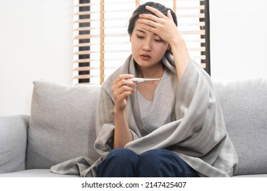 Sick, influenza asian young woman, girl headache have a fever, flu and check thermometer measure body temperature, feel illness sitting on sofa bed at home. Health care person on virus, covid-19. - Shutterstock ID 2147425407