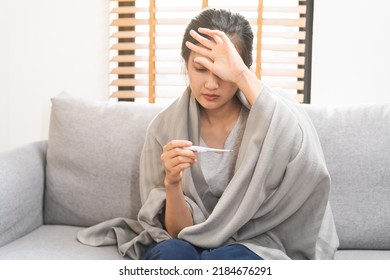 Sick, influenza asian young woman, girl headache have a fever, flu and check thermometer measure body temperature, feel illness sitting on sofa bed at home. Health care person on virus, covid-19. - Shutterstock ID 2184676291