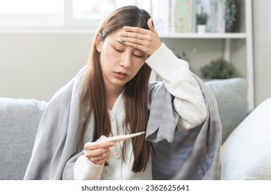 Sick, influenza asian young woman, girl have a fever, hand holding thermometer, touching forehead for check measure body temperature, illness while lying rest on sofa at home. Health care concept. - Shutterstock ID 2362625641
