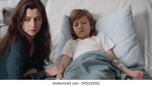 Sick Kid Boy With Lying In Bed At Hospital And Mother Holding His Hand. Young Upset Woman Sitting At Bedside Of Ill Preteen Son Resting In Hospital Bed With Nasal Breathing Tube