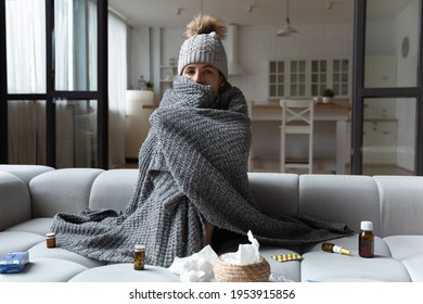 Sick Latin Female Feel Bad Ill Has Health Problems In Cold Freezing Flat With Broken Central Heating. Young Woman Sit On Sofa In Cap Covered With Knitted Plaid Taking Medicine For Fever Flu Influenza