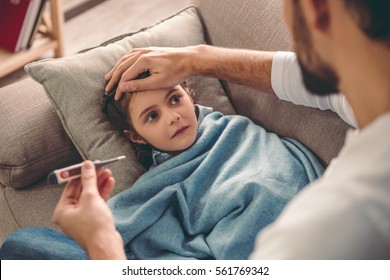 Sick little girl covered in blanket is lying on couch while her father is taking her temperature - Shutterstock ID 561769342
