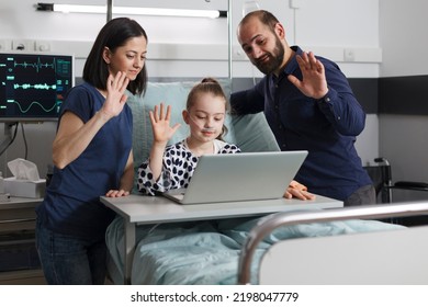 Sick Little Girl Sitting With Parents On Patient Bed While Waving At Laptop Video Call Inside Hospital Pediatrics Ward. Ill Kid In Healthcare Clinic Talking With Relatives On Virtual Online Call.