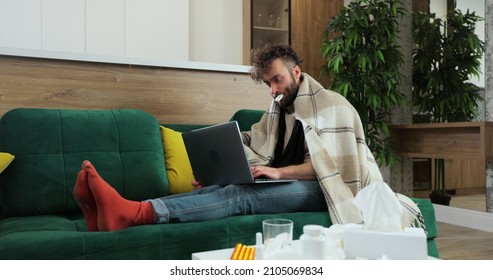 Sick male freelancer sitting on sofa at home office under plaid, working remotely, using paper tissue with termometr in mouth. Man feeling unwell, sick with running nose, need a rest. - Shutterstock ID 2105069834