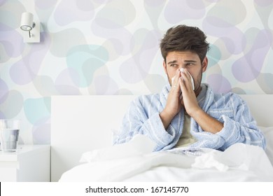 Sick Man Blowing His Nose While Sitting On Bed At Home