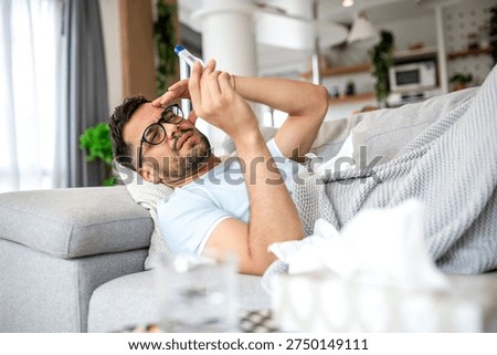 Sick man checking high temperature with thermometer on sofa. Ill male lying under blanket with fever and headache, Healthcare And Medicine concept. Young guy suffering from flu or cold symptoms home