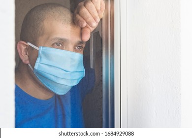 Sick Man With Face Mask Looking Out The Window Being Quarantined At Home. He Is Sad That He Cannot Get Out Due To The Covid-19 Coronavirus Pandemic.Space For Text.