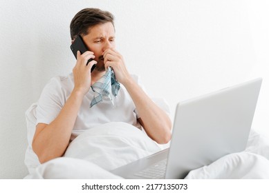 Sick Man With Flu Symptoms Using Mobile Phone And Laptop While Sitting On Bed In Bedroom. Sad Ill Guy Holding Handkerchief And Calling On Smartphone. Remote Work On Sick Leave, Cyberchondria Concept.