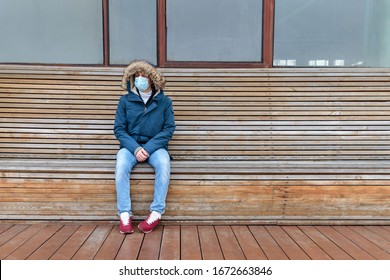 Sick Man With Hood Sitting Alone On Bench, Wearing Facial Mask Against Transmissible Infectious Diseases, Covid-19. Empty City From People Due To Coronavirus Pandemic. Life In Isolation And Quarantine