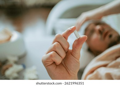 Sick man lying in bed, grasping a pill between his fingers, surrounded by used tissues and a thermometer resting on the bedside table, reflecting illness and discomfort