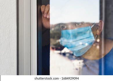 Sick Man With Mask Is Looking At The Landscape Through The Window Because He Is In Quarantine And Cannot Leave Home Due To The Covid-19 Coronavirus Pandemic.