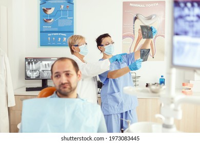 Sick Man Patient Waiting For Teeth Examination Sitting On Dental Chair In Stomatology Clinic Room. Stomatologist Doctor And Medical Nurse Preparing For Orthodontic Surgery While Examining Radiography