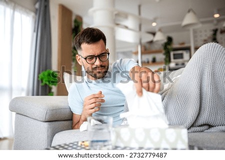 Sick Man Reaching for Tissue Box, Flu and Cold Season Concept. Ill Male with Glasses Feeling Unwell and Lying Under Blanket. Healthcare and Medicine Concept of Viral Infection and Nasal Congestion.