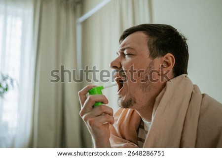 Sick man is sitting on a bed, covered with a blanket, and using a throat spray to relieve his sore throat, demonstrating the concept of home treatment for illness