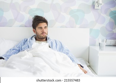 Sick Man With Thermometer In Mouth Reclining On Bed At Home