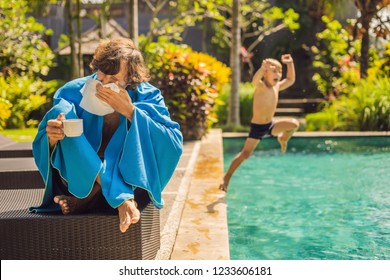 Sick Man Traveler. The Man Caught A Cold On Vacation, Sits Sad At The Pool Drinking Tea And Blows His Nose Into A Napkin. His Son Is Healthy And Swimming In The Pool. Travel Insurance Concept