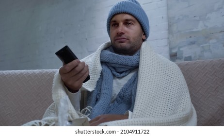 Sick Man In Warm Beanie, Scarf And Blanket Clicking Channels While Watching Tv